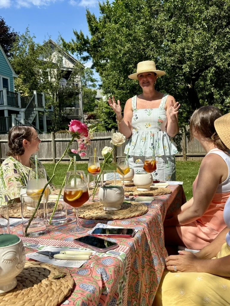 A woman wearing a wide-brimmed straw hat and a sleeveless, floral dress is speaking and gesturing with her hands at an outdoor gathering. She is standing at a table with several seated women, floral centerpieces, drinks, and a colorful tablecloth, in a backyard with green trees and houses.