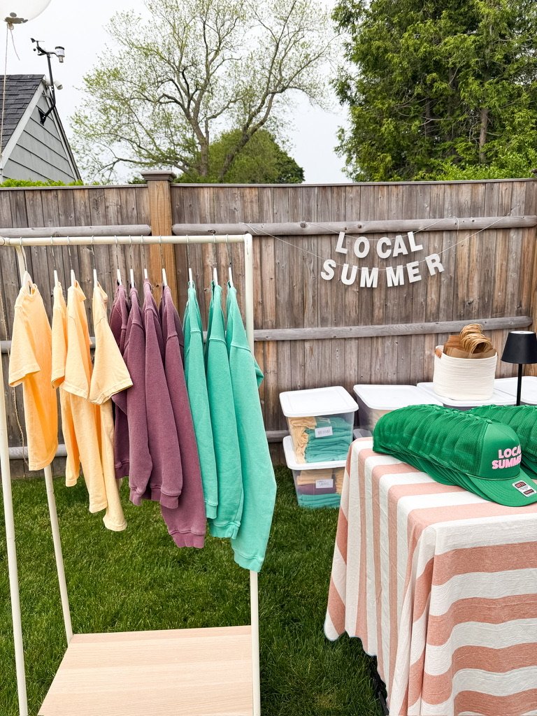 A small outdoor booth with pastel-colored T-shirts and hoodies hanging on a rack, a table with a striped tablecloth displaying a green hat with 'Local Summer' branding, and a wooden fence with a sign that reads 'Local Summer' in white letters, surrounded by trees and grass.