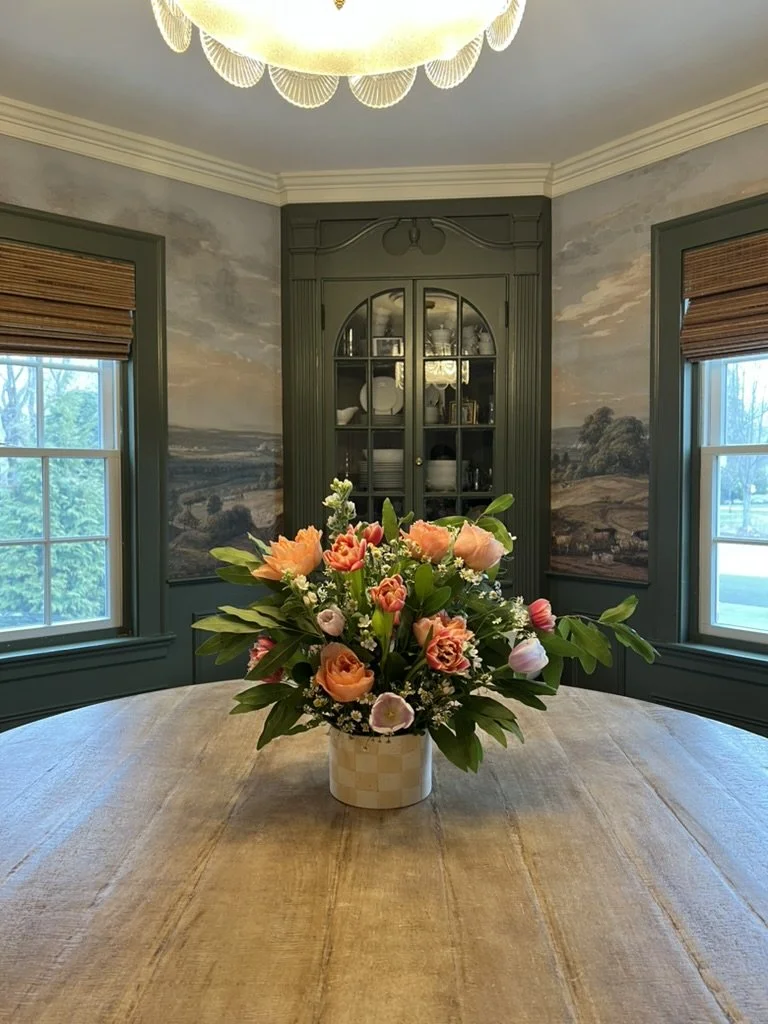 A vase of assorted pink, peach, and white flowers on a wooden dining table in a room with green trim and a landscape mural. Two windows with brown blinds are on either side of a dark green cabinet filled with dishes, and a ceiling light fixture is above.