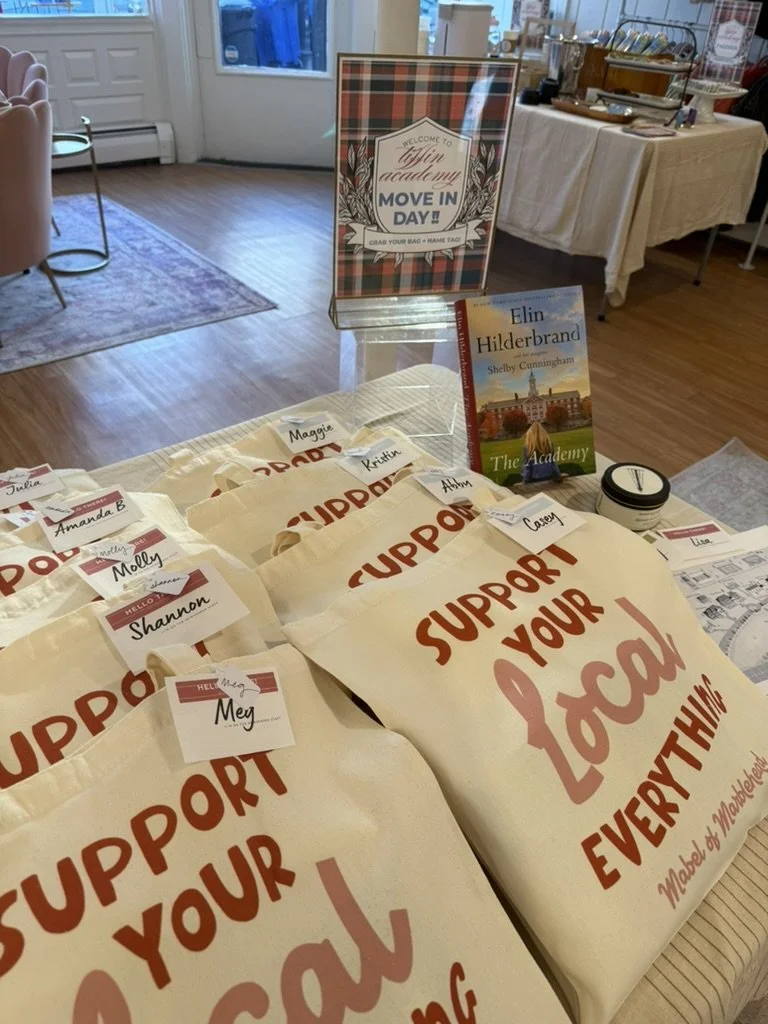 Support your local event tote bags with name tags on a table at a community event, with a sign welcoming attendees to a move-in day at Elin Hilderbrand's book giveaway, and a copy of her book 'The Hotel Nantucket' on display.