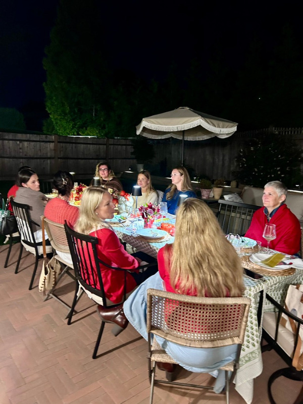 Group of nine women and one man gathered around a table for a dinner party outside at night, under a large umbrella, with a fence and trees in the background.
