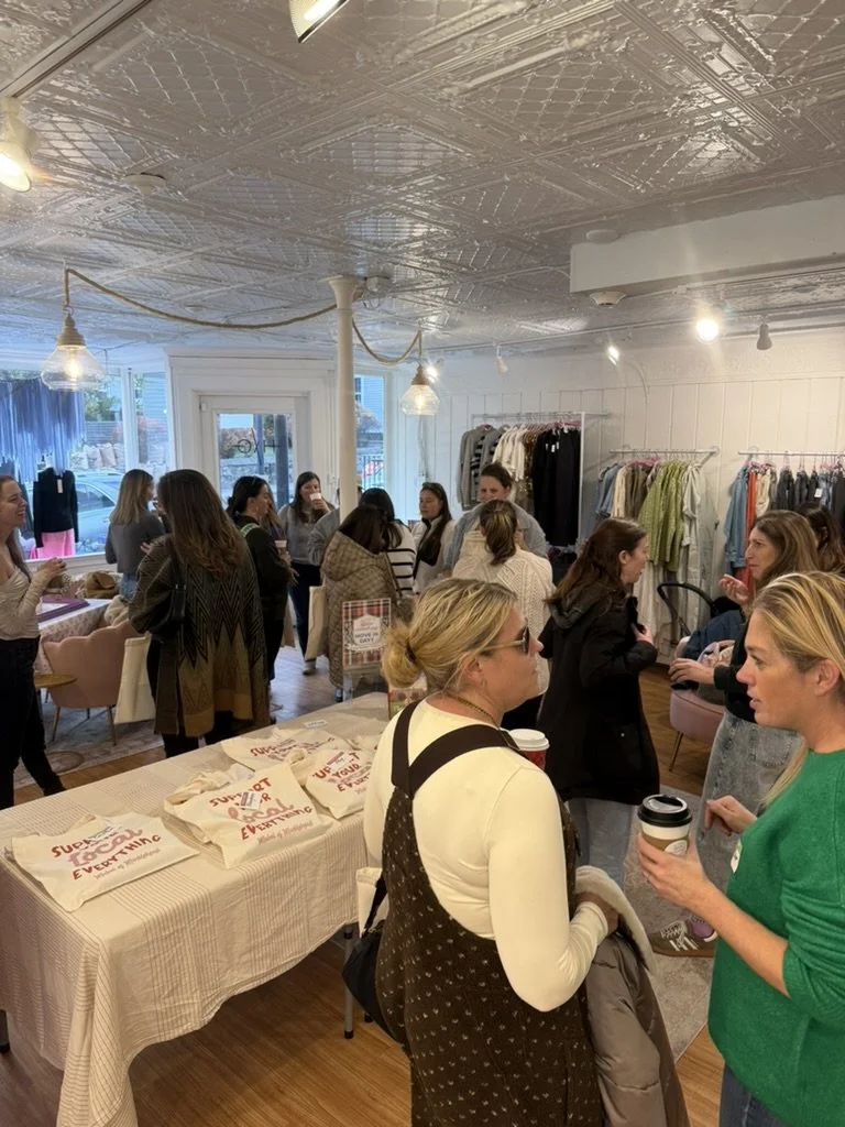 Women gathered in a retail clothing store for an event or sale, with tables displaying totes and racks of clothing in the background.