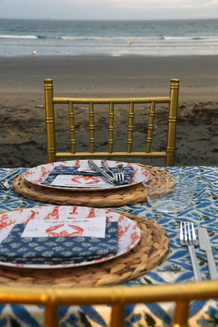 Beachside dining setup with two plates, utensils, and glasses on a patterned tablecloth, with the ocean and sky in the background.