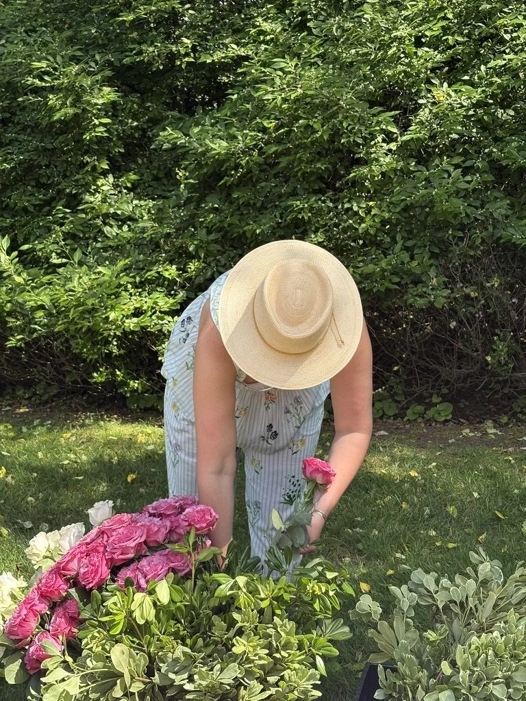 Person wearing a large sun hat and striped dress, tending to pink and white peonies in a garden surrounded by green foliage.