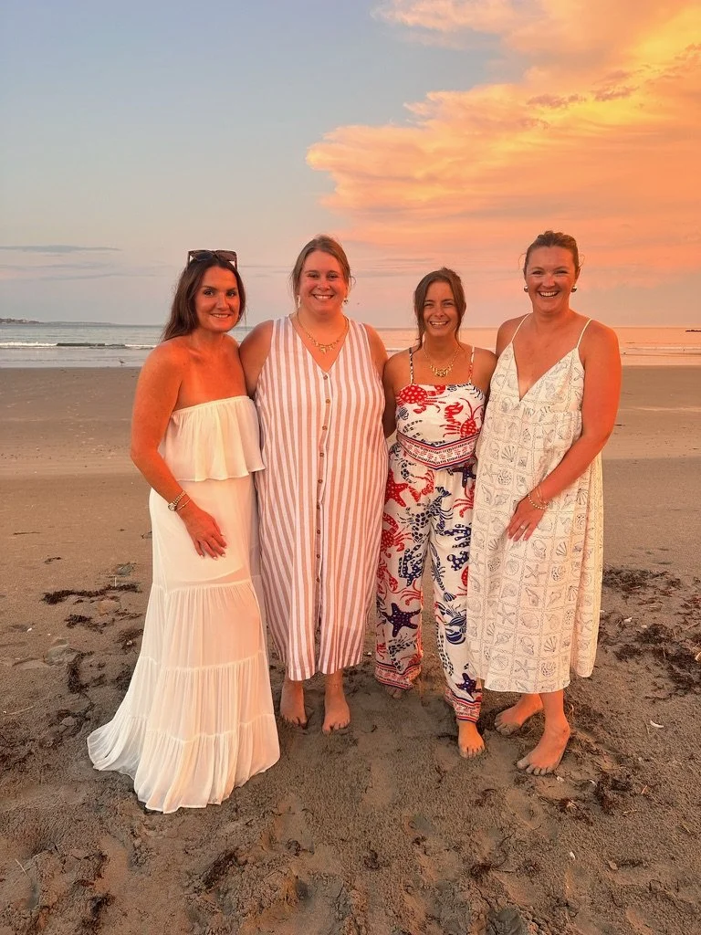 Four women standing together on a beach during sunset, smiling, wearing summer dresses and casual outfits.
