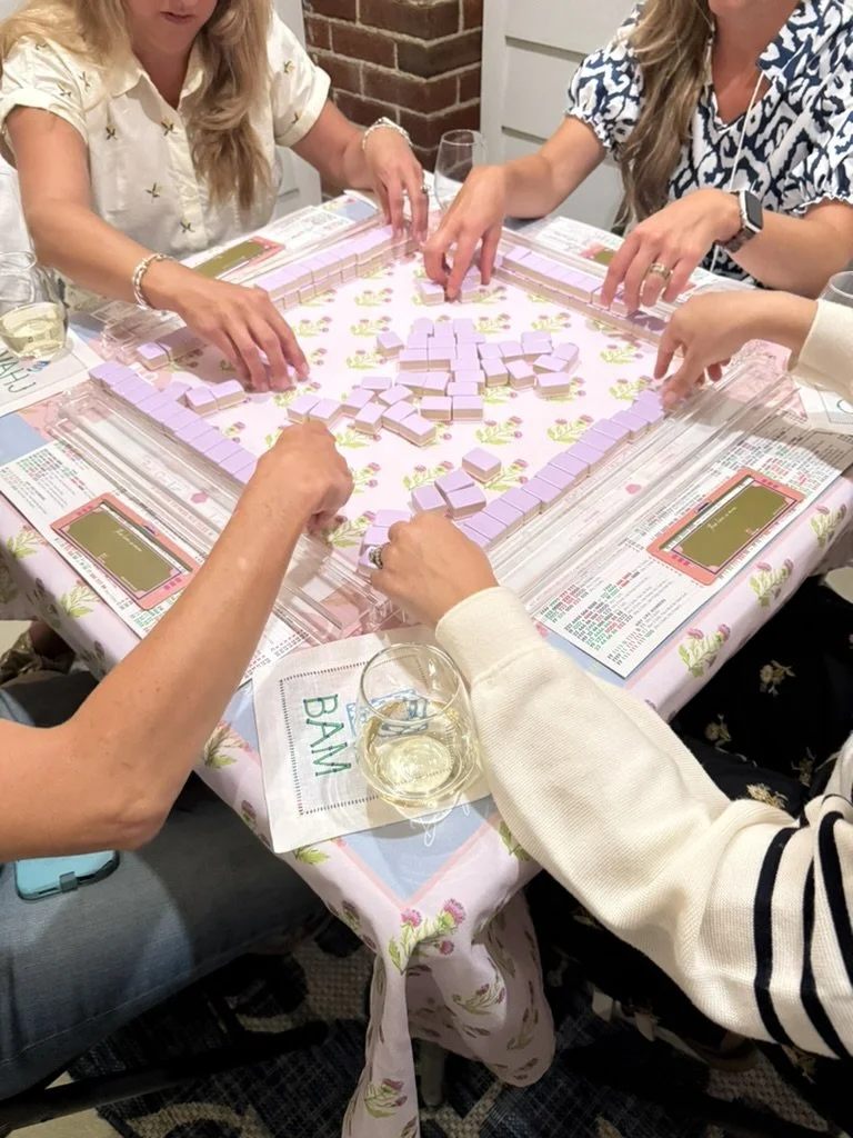 People playing mahjong at a round table covered with a floral tablecloth, with drinks and printed game instructions nearby.