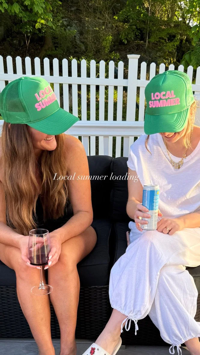 Two women wearing green hats with pink text that says 'Local Summer' sitting outdoors with drinks, a wine glass and a canned beverage, on a patio with a white picket fence and trees in the background.
