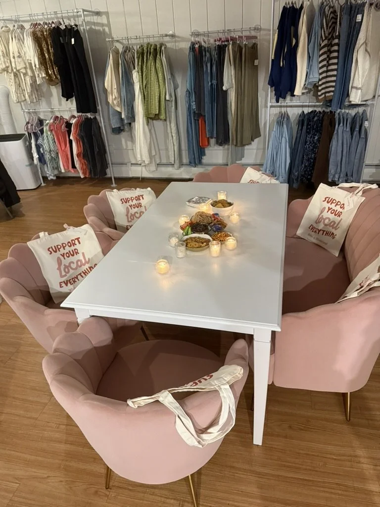 A dining table surrounded by pink upholstered chairs with support your local business bags on the chairs, on a wooden floor, in a clothing store with racks of clothes in the background, and candles and bowls on the table.