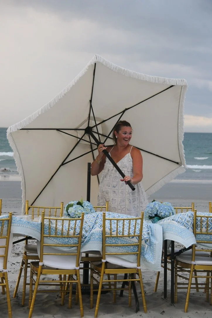 A woman in a white dress holding a large white beach umbrella, standing behind a table with blue and white patterned tablecloths and floral centerpieces on a beach.