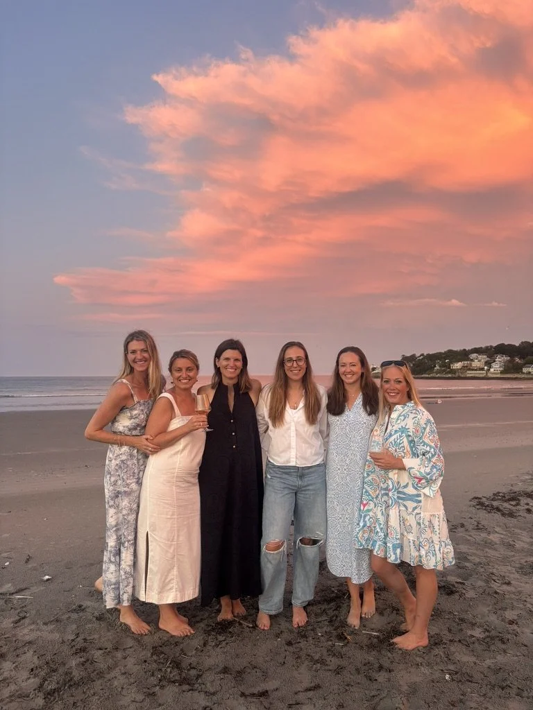 Six women standing barefoot on the beach at sunset, smiling, with a sky filled with pink and orange clouds, ocean waves, and hills with houses in the background.