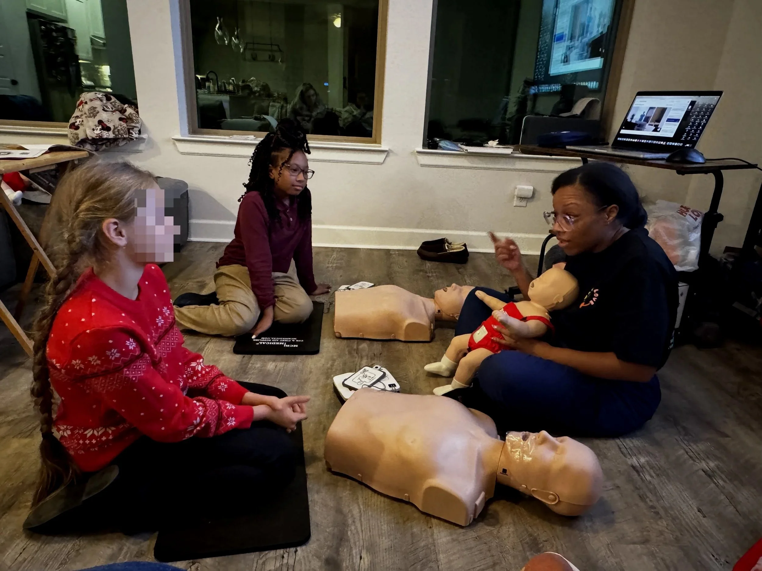 A woman demonstrates CPR on a mannequin while two children watch, in a room with medical equipment and computers.
