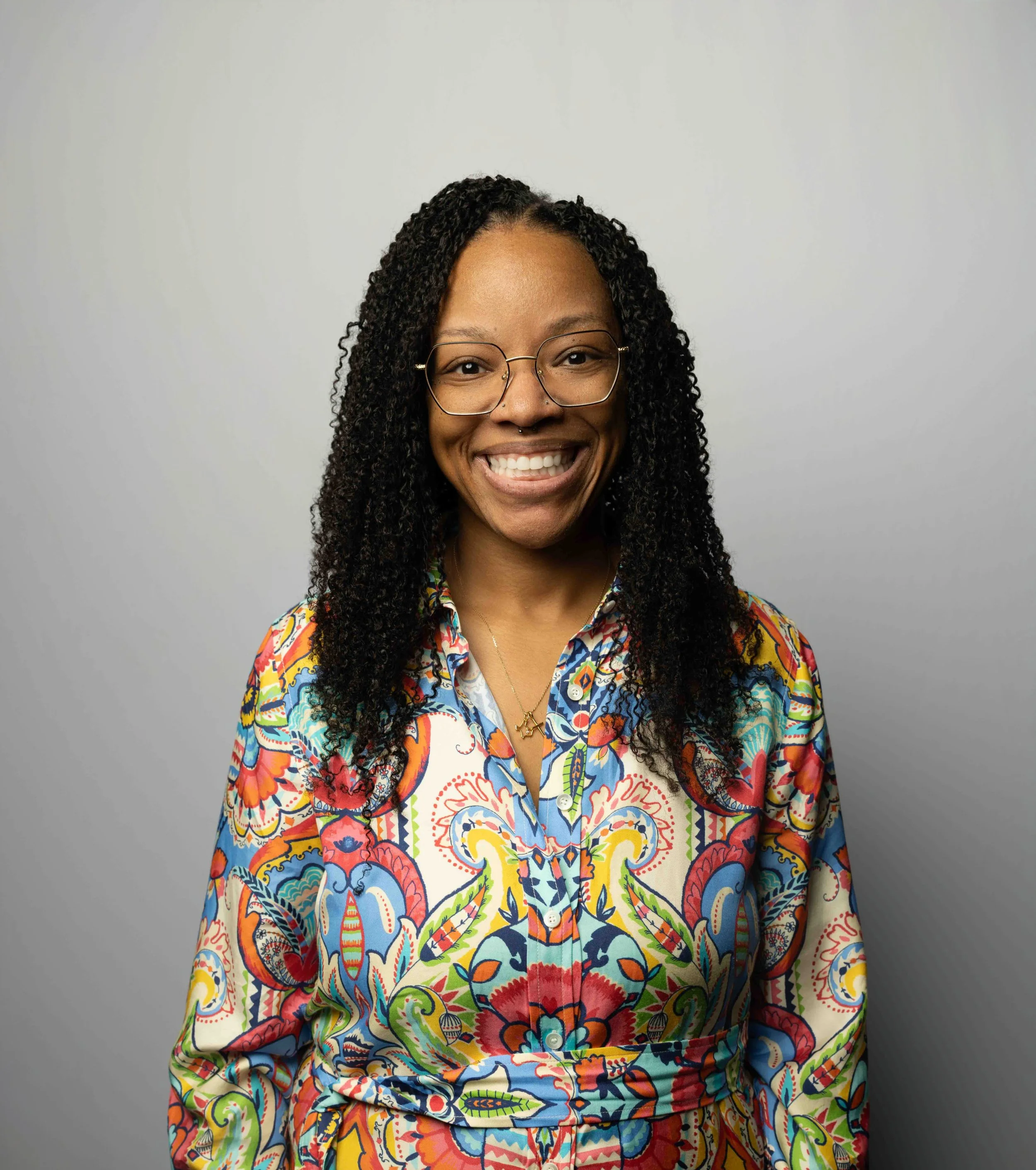 Portrait of a smiling woman with curly hair wearing glasses and a colorful, patterned blouse against a plain light gray background.