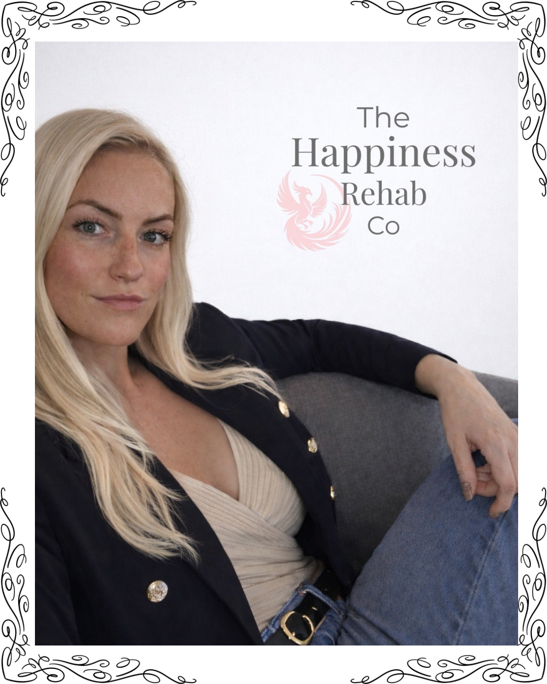 A woman with long blonde hair, wearing a black blazer with gold buttons, a beige top, and jeans, sitting on a gray couch with her arm resting on the back, in front of a white wall with a logo for The Happiness Rehab Co.