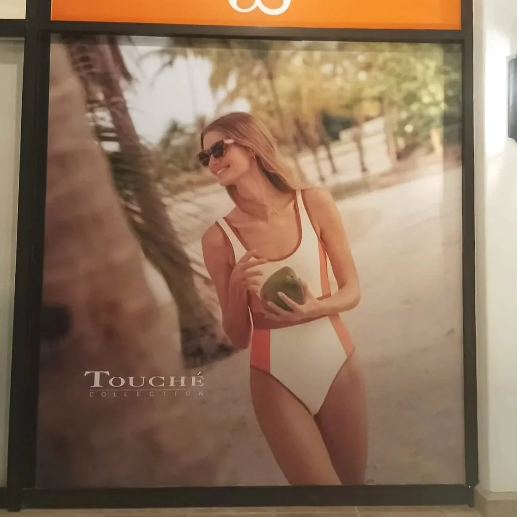 Una mujer con gafas de sol, vestido de baño de una pieza, en una playa tropical, sosteniendo un coco y sonriendo.