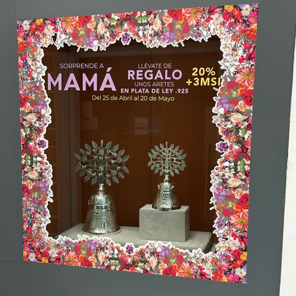 Cartel publicitario con un marco de flores coloridas que promociona una oferta de regalos para mamá, con detalles en letras moradas, amarillas y blancas. Muestra dos relojes de plata en exhibición en el centro.