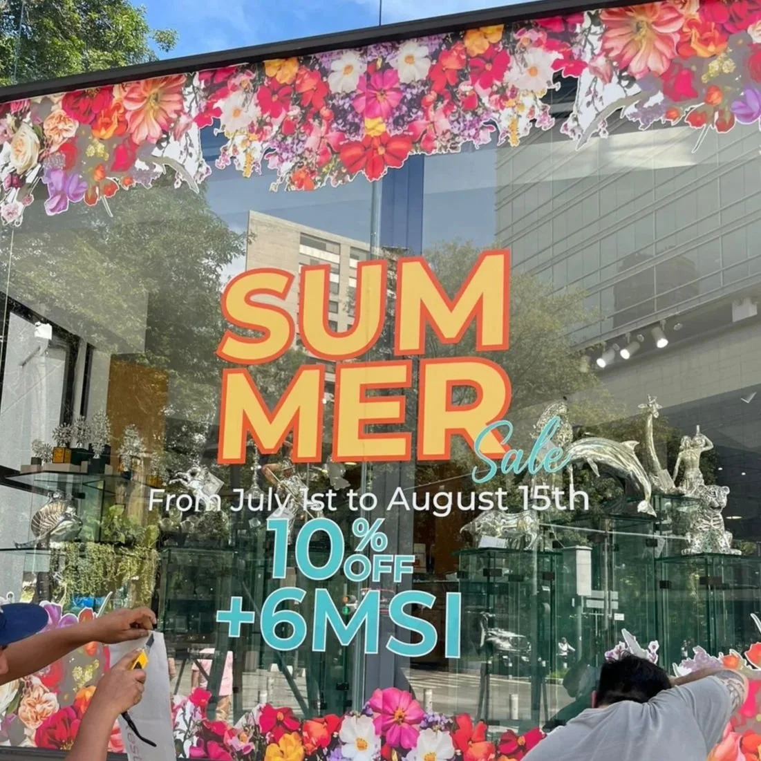 Anuncio de venta de verano en una tienda con decoraciones florales. Texto promocional en el vidrio que dice: 'SUMMER Sale, del 1 de julio al 15 de agosto, 10% de descuento + 6 MSI'.