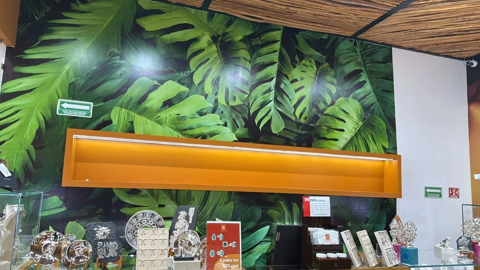 Mural con hojas grandes de plantas tropicales en tonos verdes en la pared. Mostrador con objetos decorativos de metal y artículos en exhibición en un establecimiento.