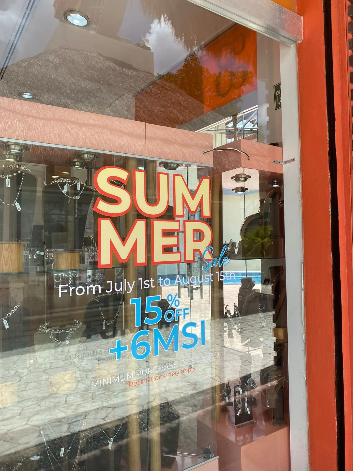 Cartel de promoción de verano en una tienda que ofrece un 15% de descuento más 6 MSI en compras realizadas del 1 de julio al 15 de agosto, con un fondo que muestra joyas en exhibición y reflejos del exterior en la ventana