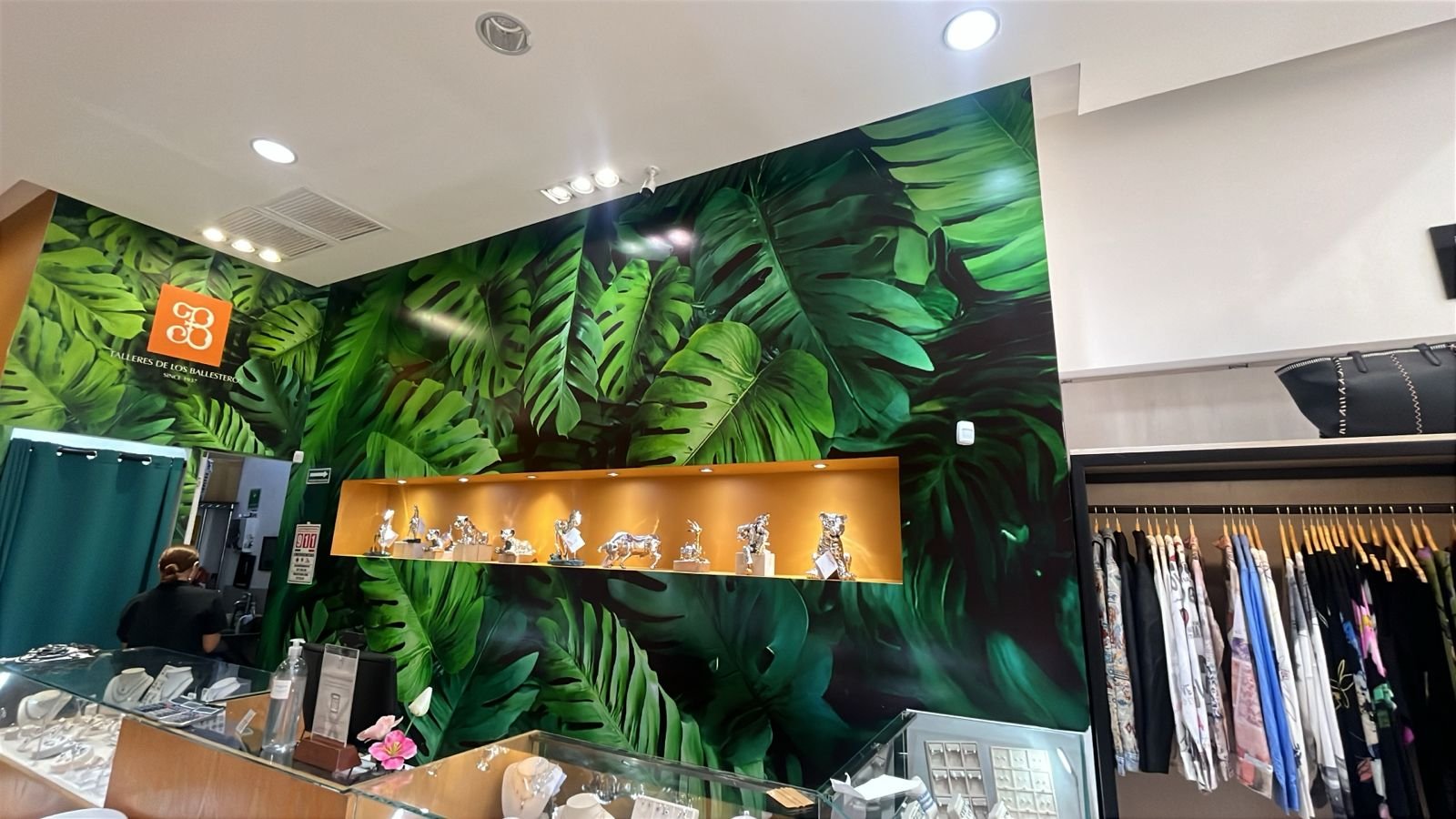 Interiores de una tienda con pared decorada con papel tapiz de hojas verdes grandes, exhibiendo figuras de animales de plata en un estante de madera y ropa colgada en una esquina.