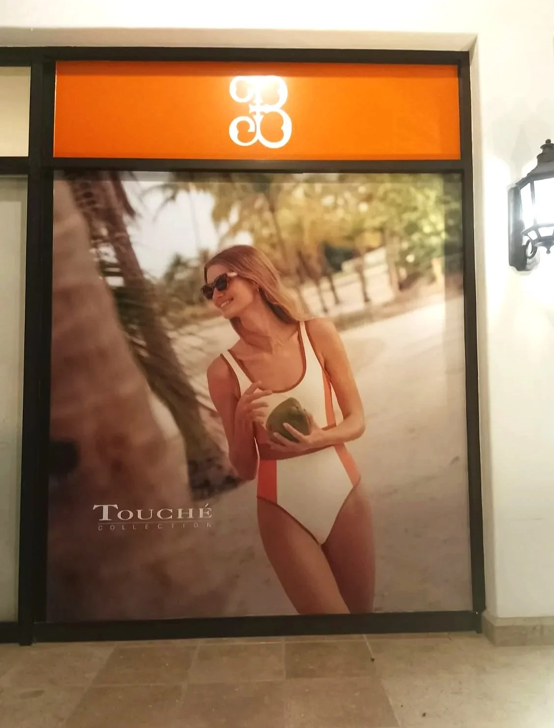 Cartel publicitario de una mujer con gafas de sol en traje de baño, en un ambiente de playa o parque, con la marca Touche Collection visible en la esquina inferior izquierda.