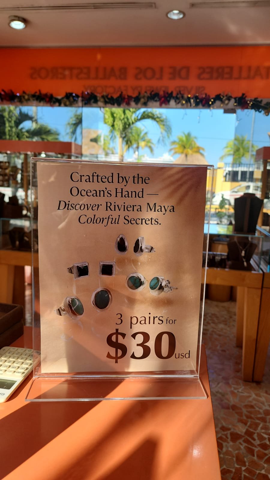 Cartel que anuncia pulseras y anillos de piedra con oferta de 3 pares por 30 dólares estadounidenses, con fondo de vista tropical y decoración colorida.