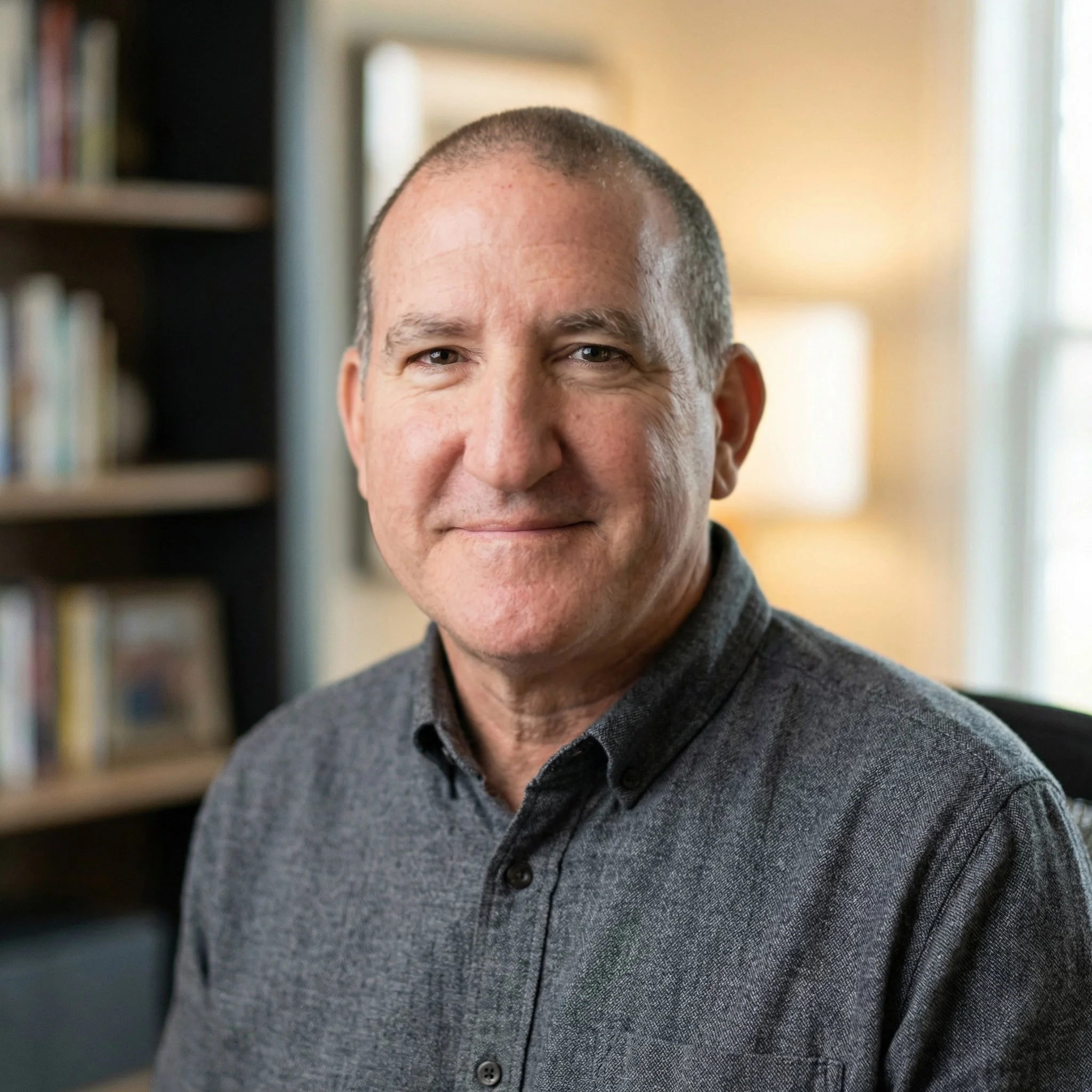 A male psychotherapist with short, dark hair and a slight smile, wearing a dark gray collared shirt, sitting in a room with a bookshelf behind him and a window to the side, natural light illuminating his face.