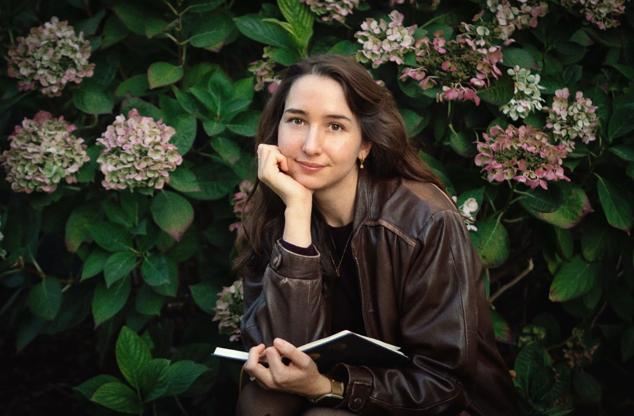 Maxine Meixner, with long dark hair, wearing a brown leather jacket, sitting outdoors in front of pink and white hydrangea flowers, resting her chin on her hand, holding a notebook.