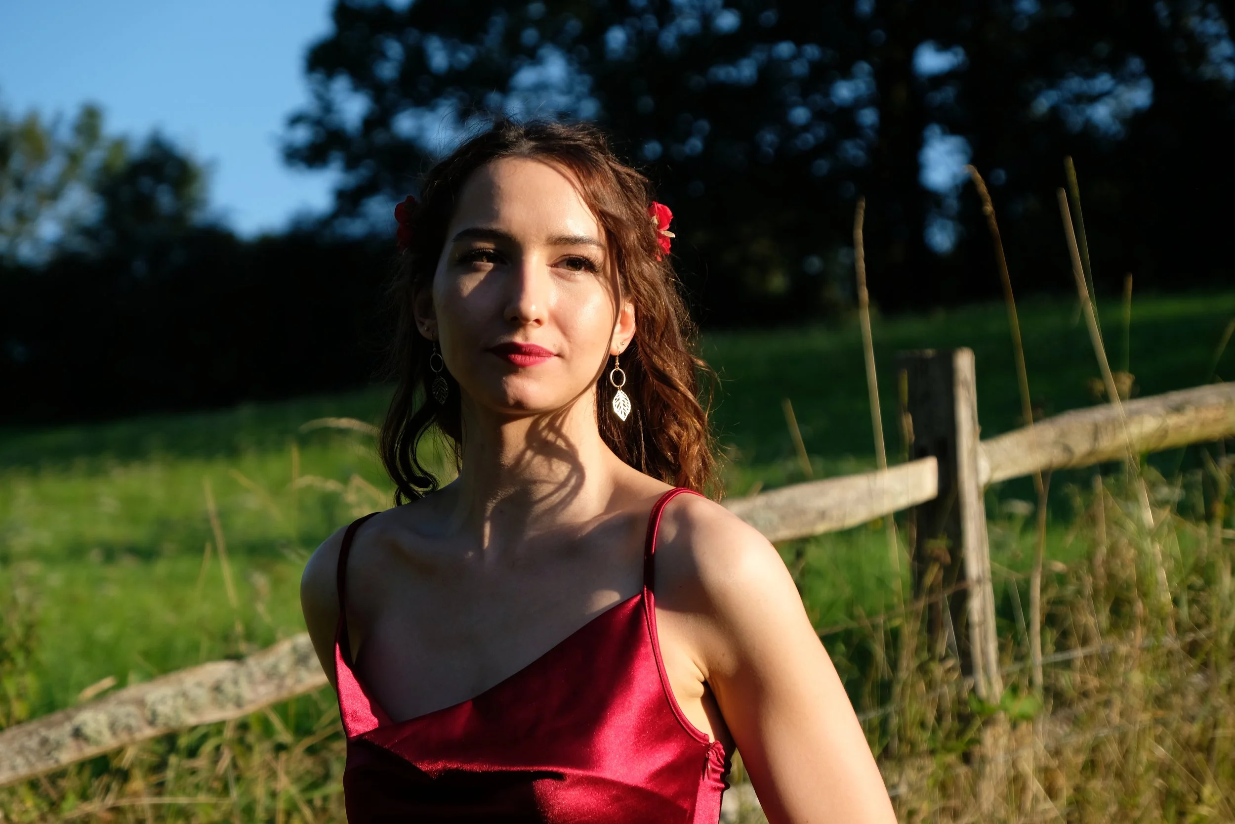 Maxine Meixner with curly brown hair wearing a red satin dress and leaf-shaped earrings, standing outdoors by a wooden fence with a grassy field and trees in the background.