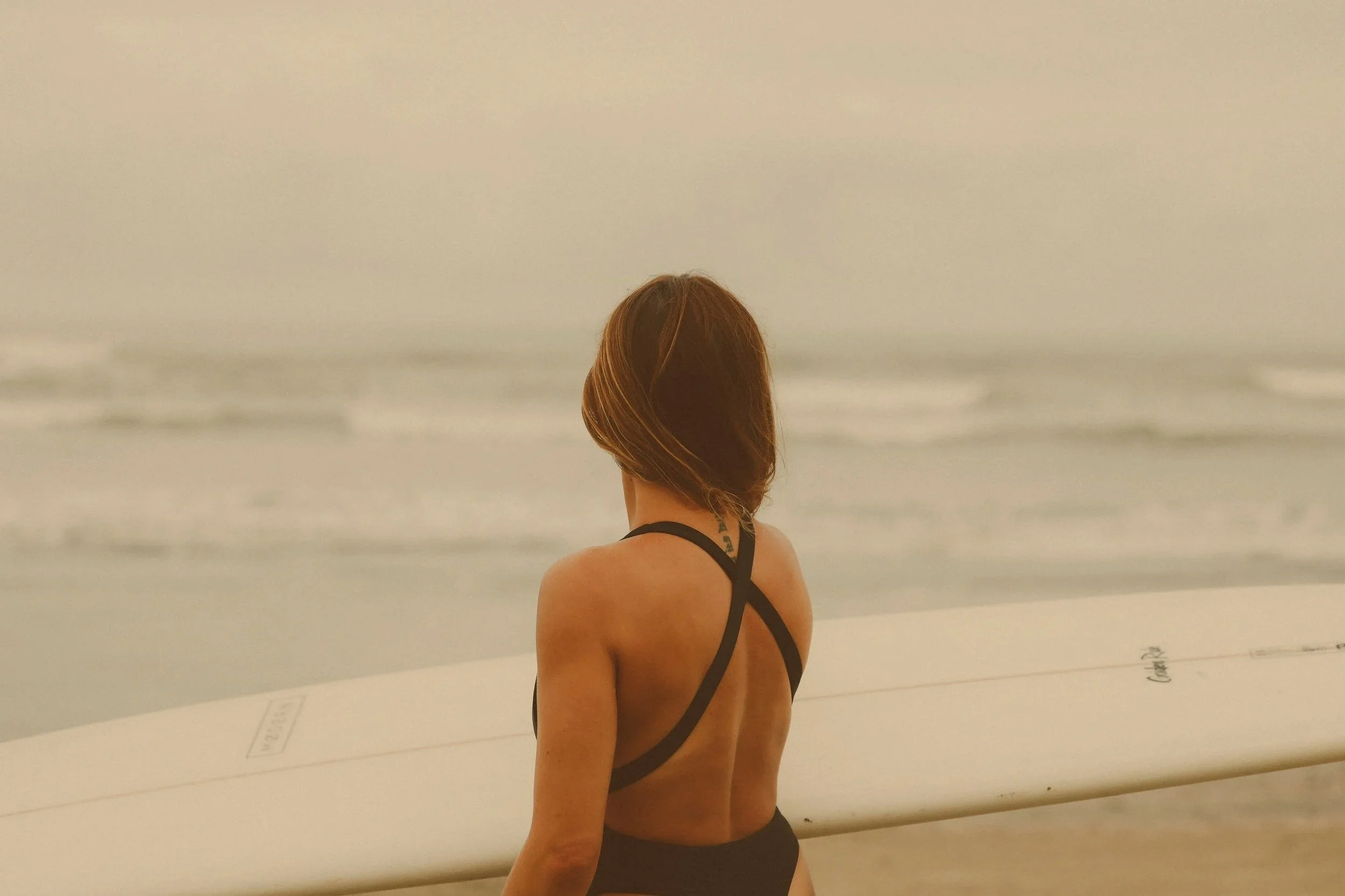 A woman with short brown hair, wearing a black swimsuit, holding a surfboard, looking towards the ocean at the beach.