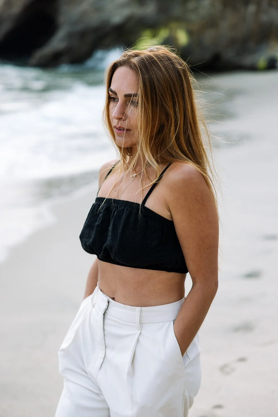 A woman with long, wavy brown hair standing on a beach with ocean waves in the background. She is wearing a black crop top and white high-waisted pants, with her hands in her pockets.