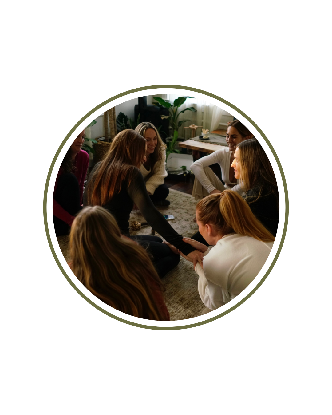 Group of women sitting on the floor in a circle, holding hands, smiling, and engaging in a group activity in a cozy living room with plants and sunlight.