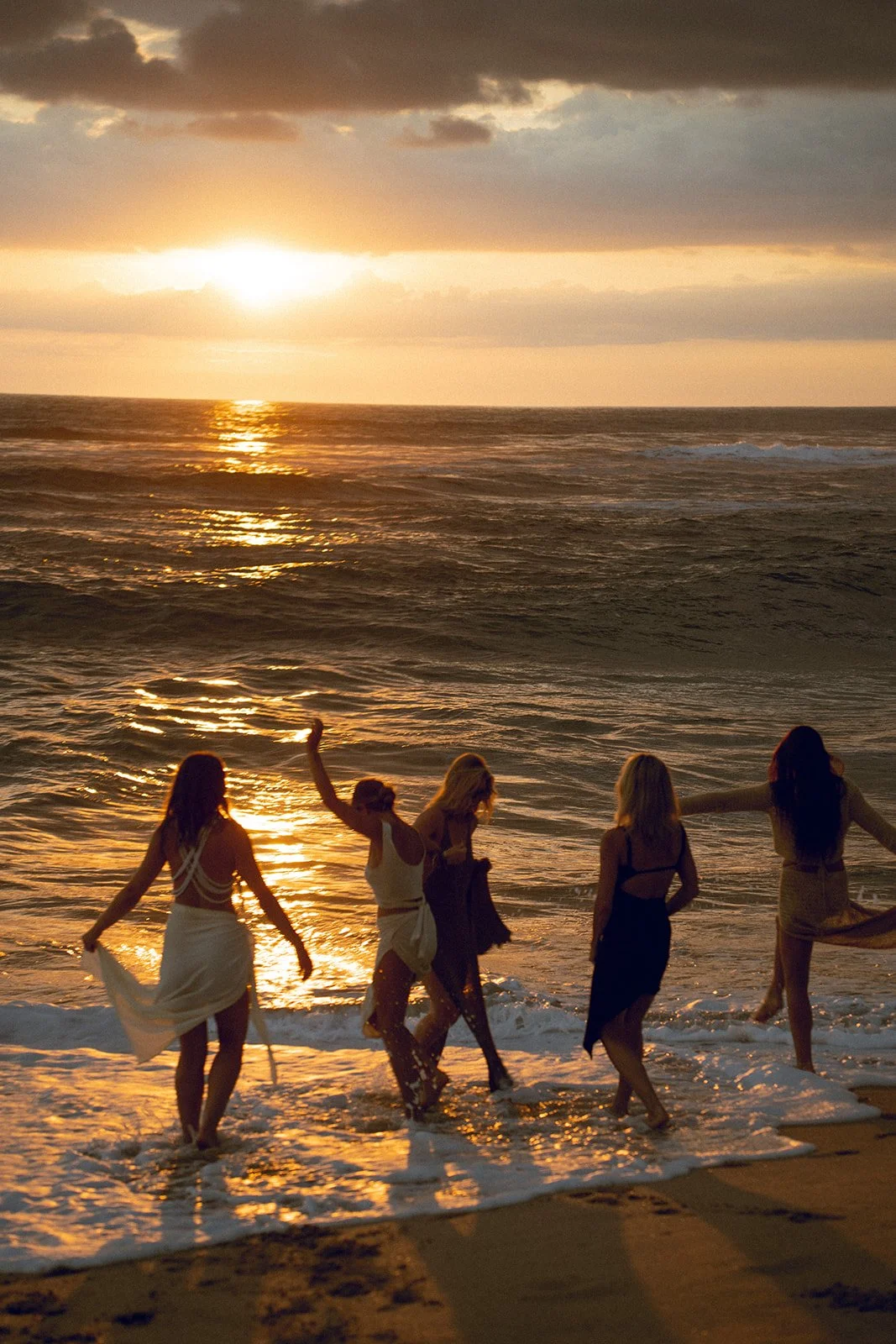 Five women are walking and playing at the shoreline of a beach during sunset, with the sun reflecting on the water and gentle waves.