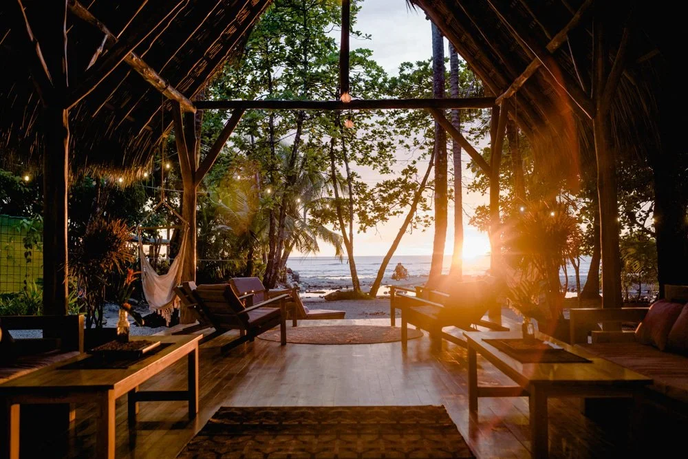 View of a beach sunset from inside a rustic open-air lounge with wooden furniture and trees around.