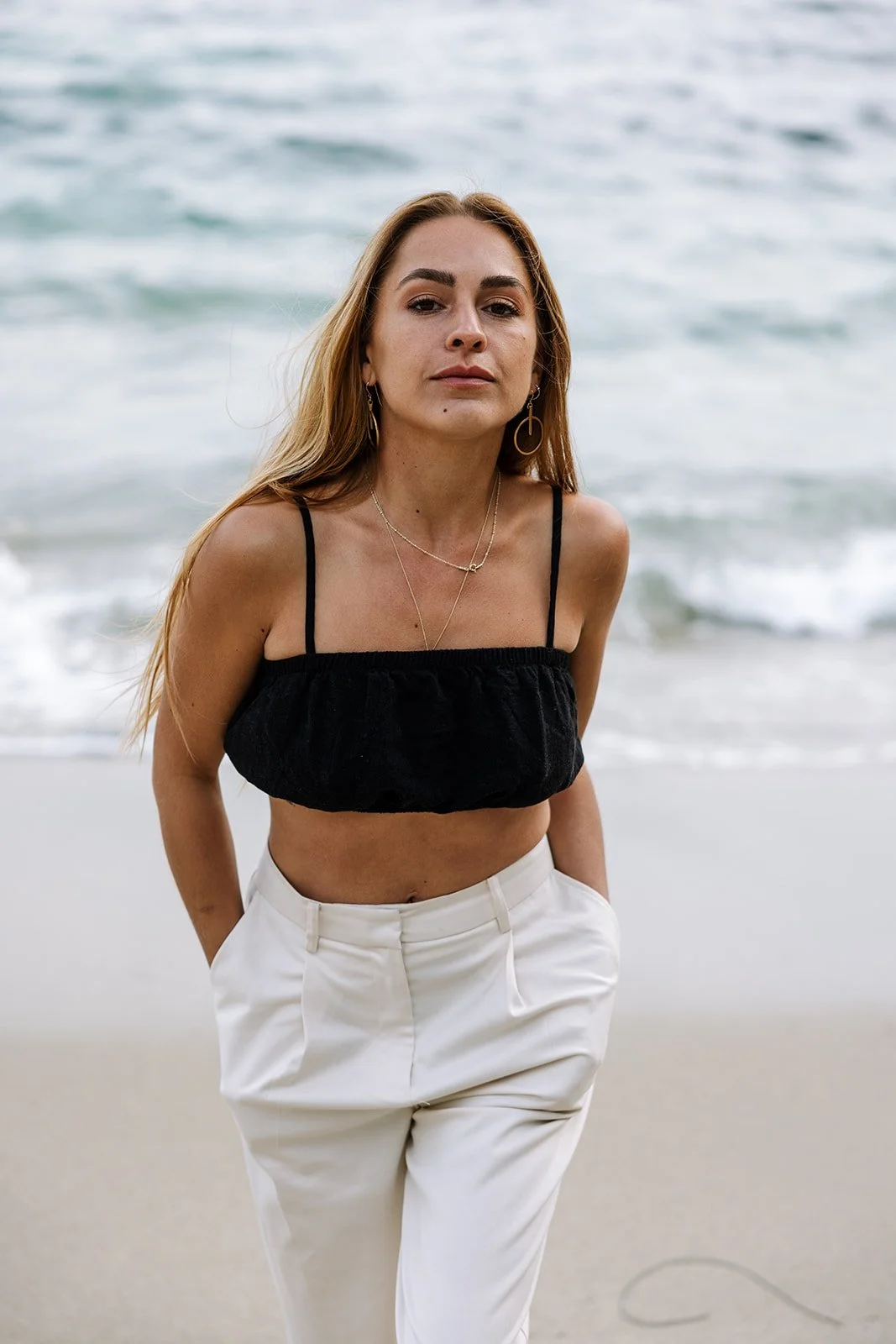 Woman with long hair standing on a sandy beach near the ocean, wearing a black crop top, white high-waisted pants, and jewelry including earrings and layered necklaces.