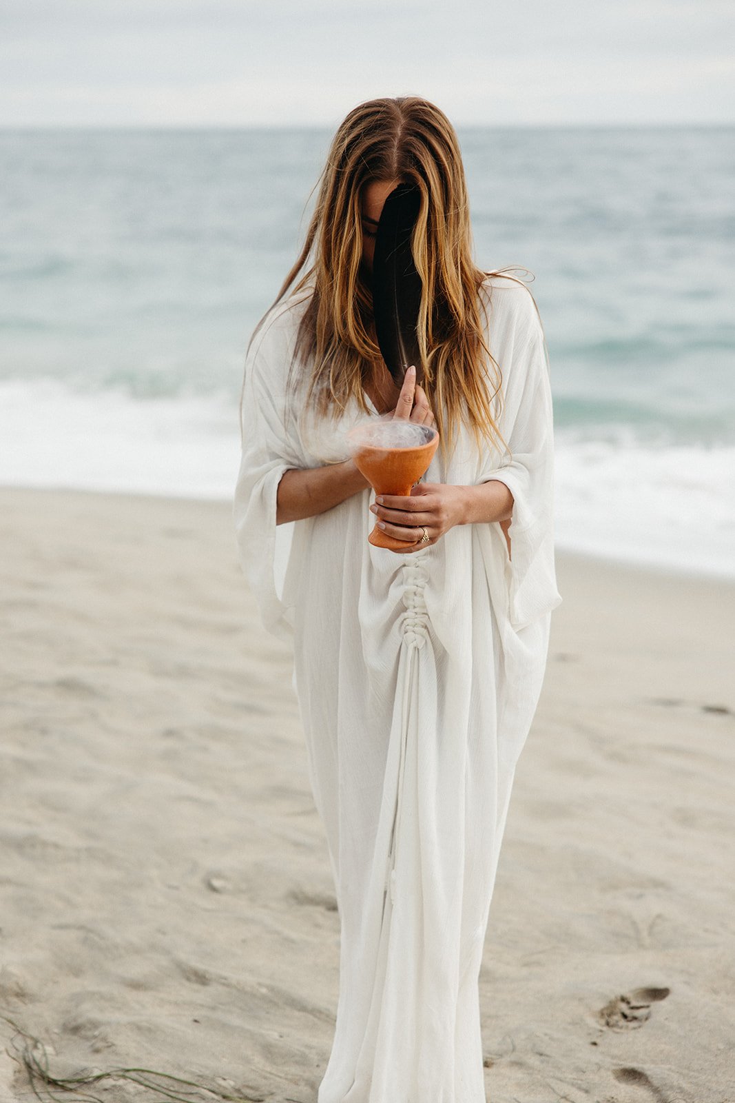 A woman with long red hair standing on a beach, holding a hookah pipe with a black feather in front of her face, dressed in a loose white dress.