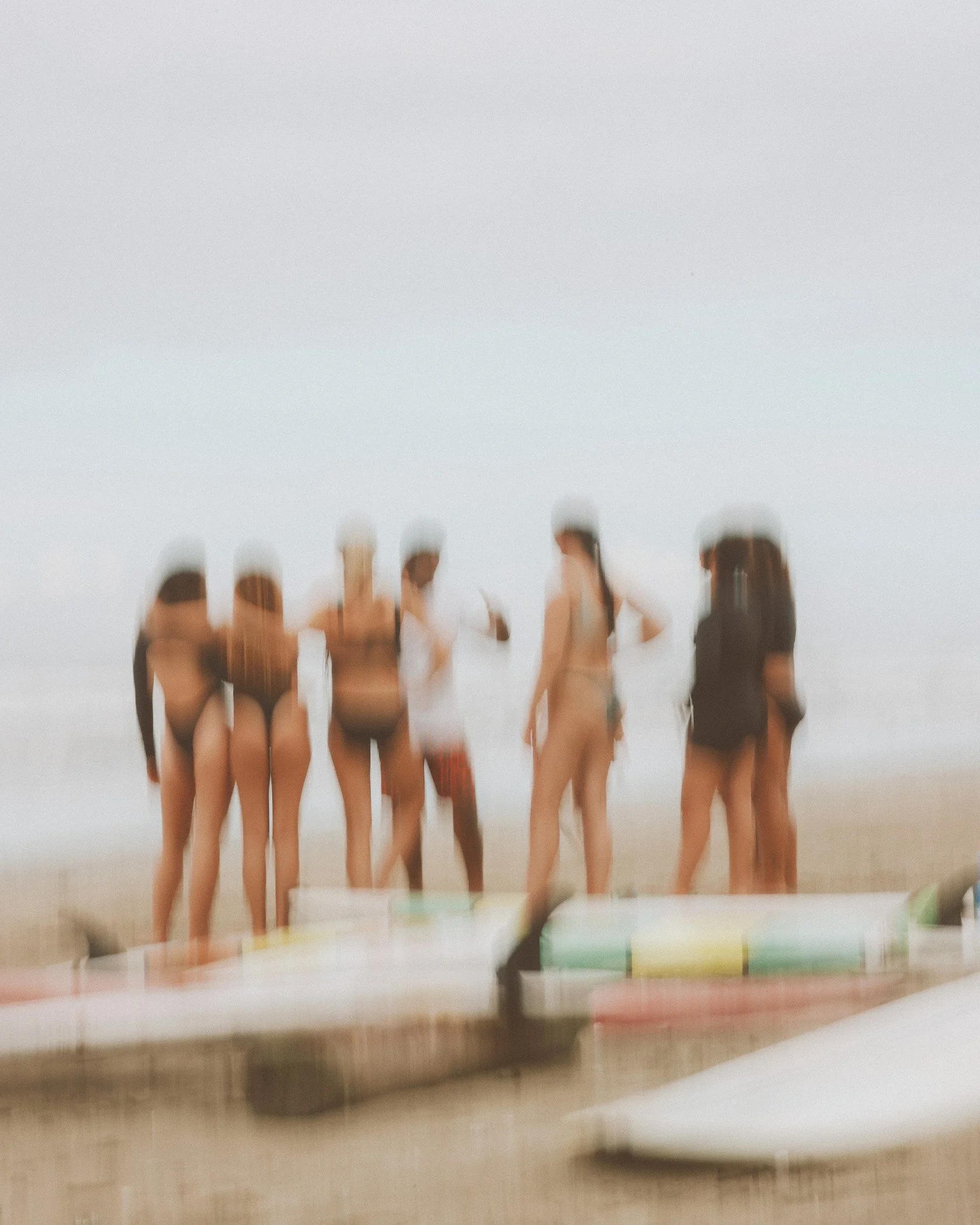 Group of people on a beach, some wearing swimsuits, some standing, some looking at their phones, with paddleboards in the foreground, under a cloudy sky.