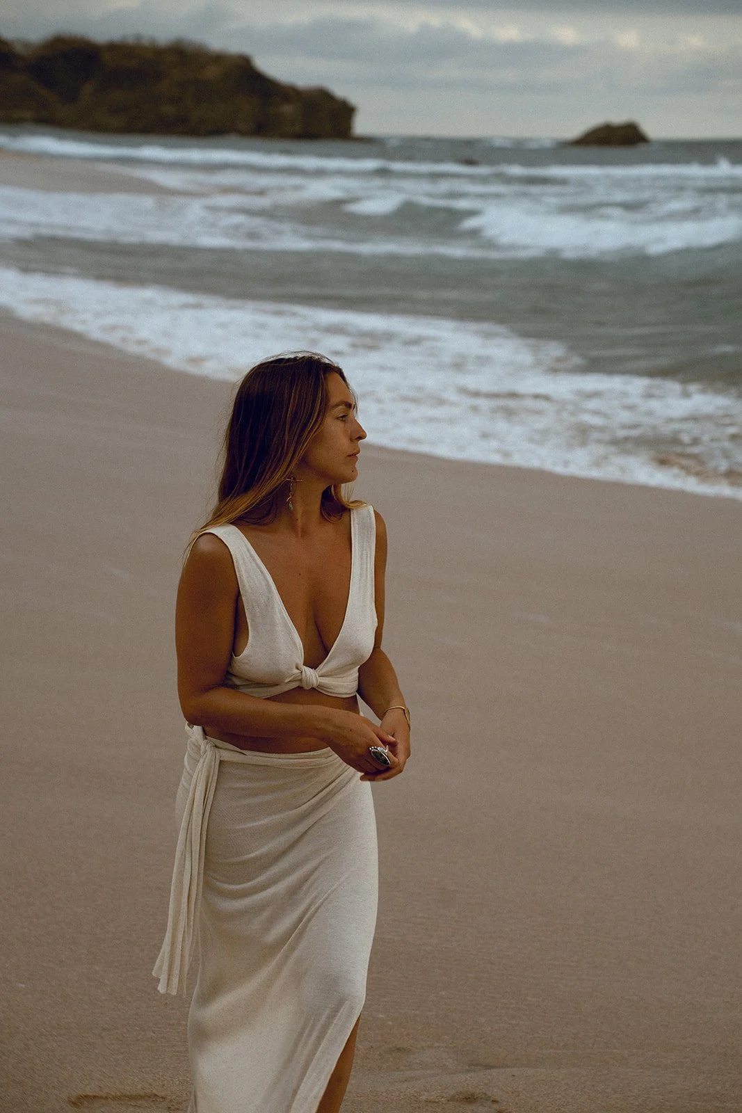 A woman in a white dress with a knot at the front stands on the sandy beach, facing ocean waves, with land and rock formations in the background, during cloudy weather.