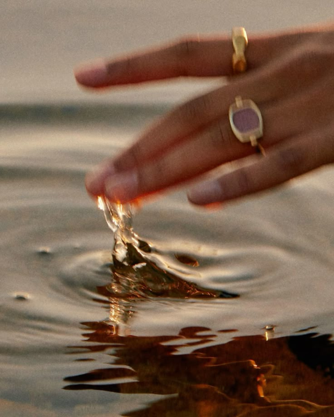 Close-up of a hand with rings reaching into water, creating ripples and splashes, during sunset or sunrise.