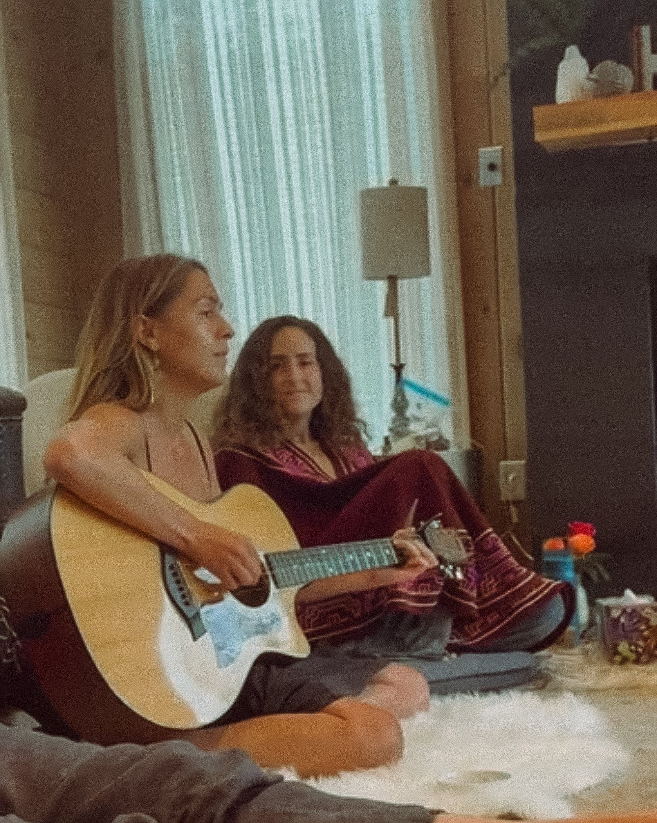 Two women sitting on a cozy rug in a room with natural light, one playing guitar and both relaxing.