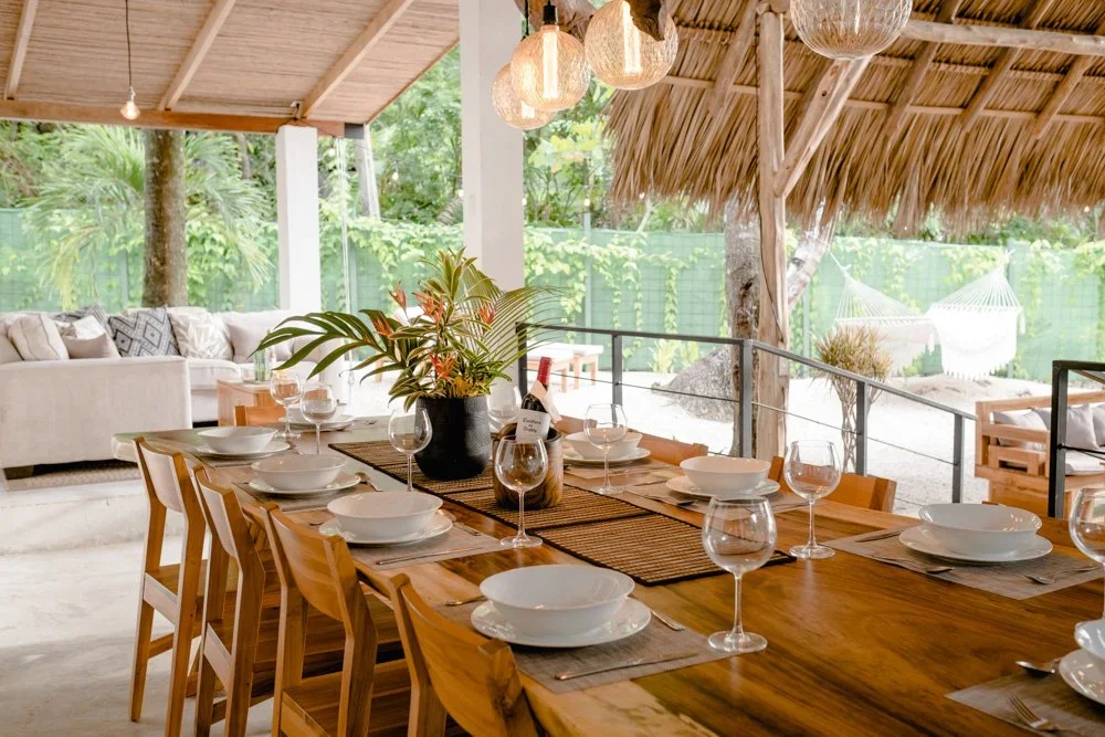 Outdoor dining area with a large wooden table set with bowls, wine glasses, and a potted plant centerpiece, surrounded by a covered patio with wood ceiling and hanging lanterns, overlooking a lush green garden with a hammock in the background.