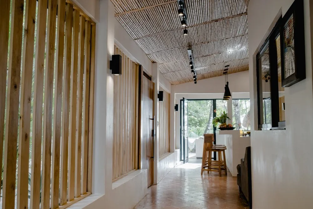 Interior of a modern home with wooden slat walls, a tiled ceiling with track lighting, an open doorway leading outside, a small kitchen area with bar stools, and a view of green trees outside.