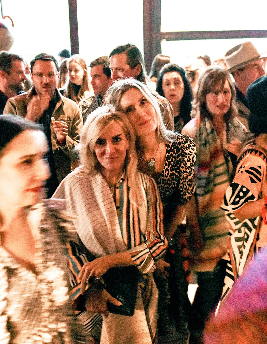 People gathered at a social event, with two women in the center posing for the camera, surrounded by others engaging or looking around, in a lively indoor setting.