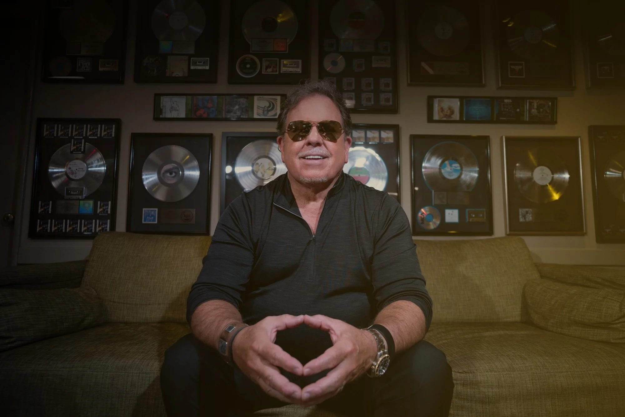 A man sitting on a sofa, wearing sunglasses and a black shirt, smiling, with framed platinum and gold records on the wall behind him.