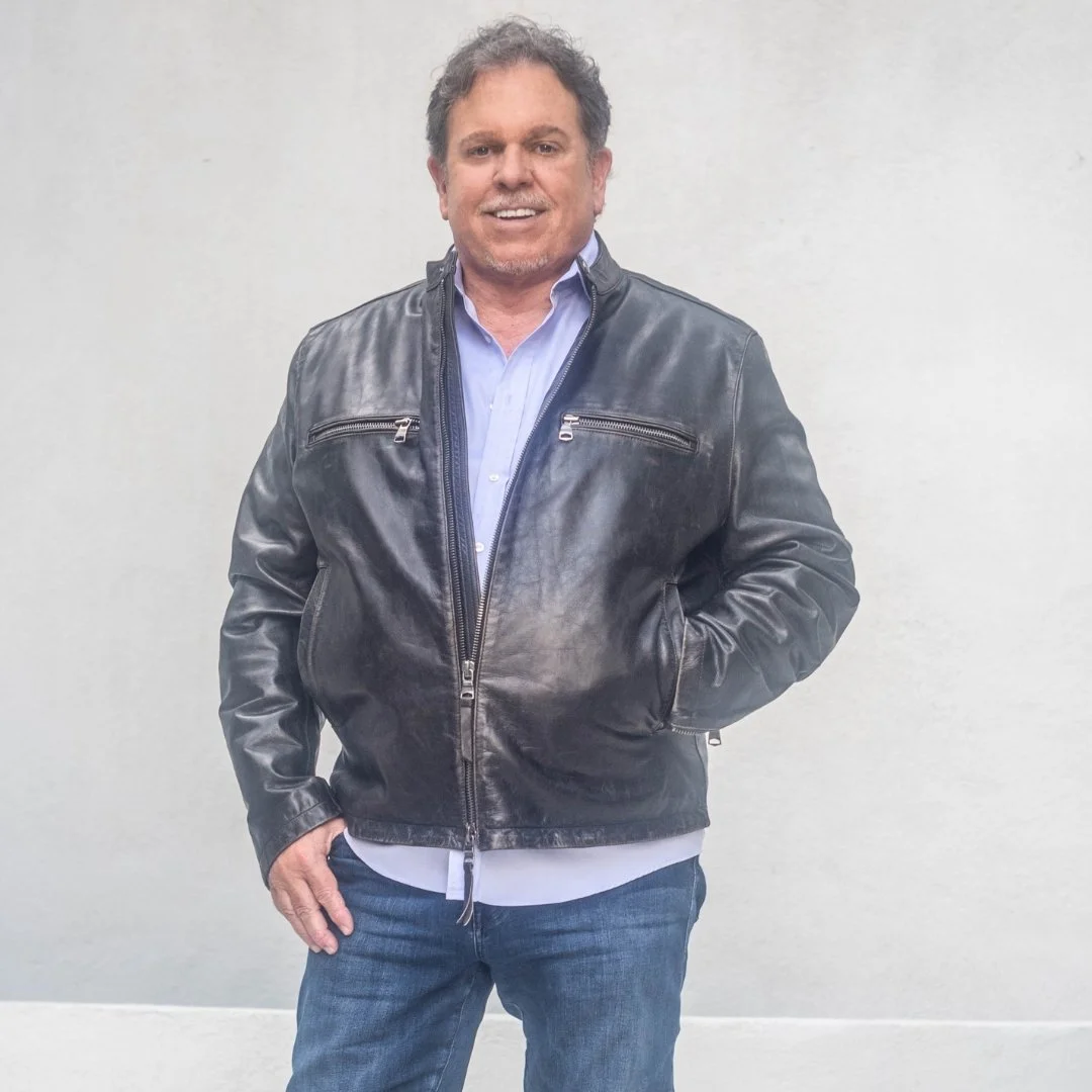 A man with curly hair wearing a black leather jacket over a light blue dress shirt, standing against a plain white background.