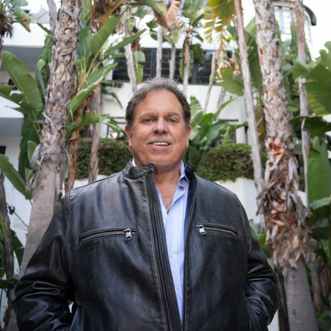 A middle-aged man with gray hair and a light beard smiling outdoors, wearing a black leather jacket over a light blue shirt, standing in front of tropical plants and trees.