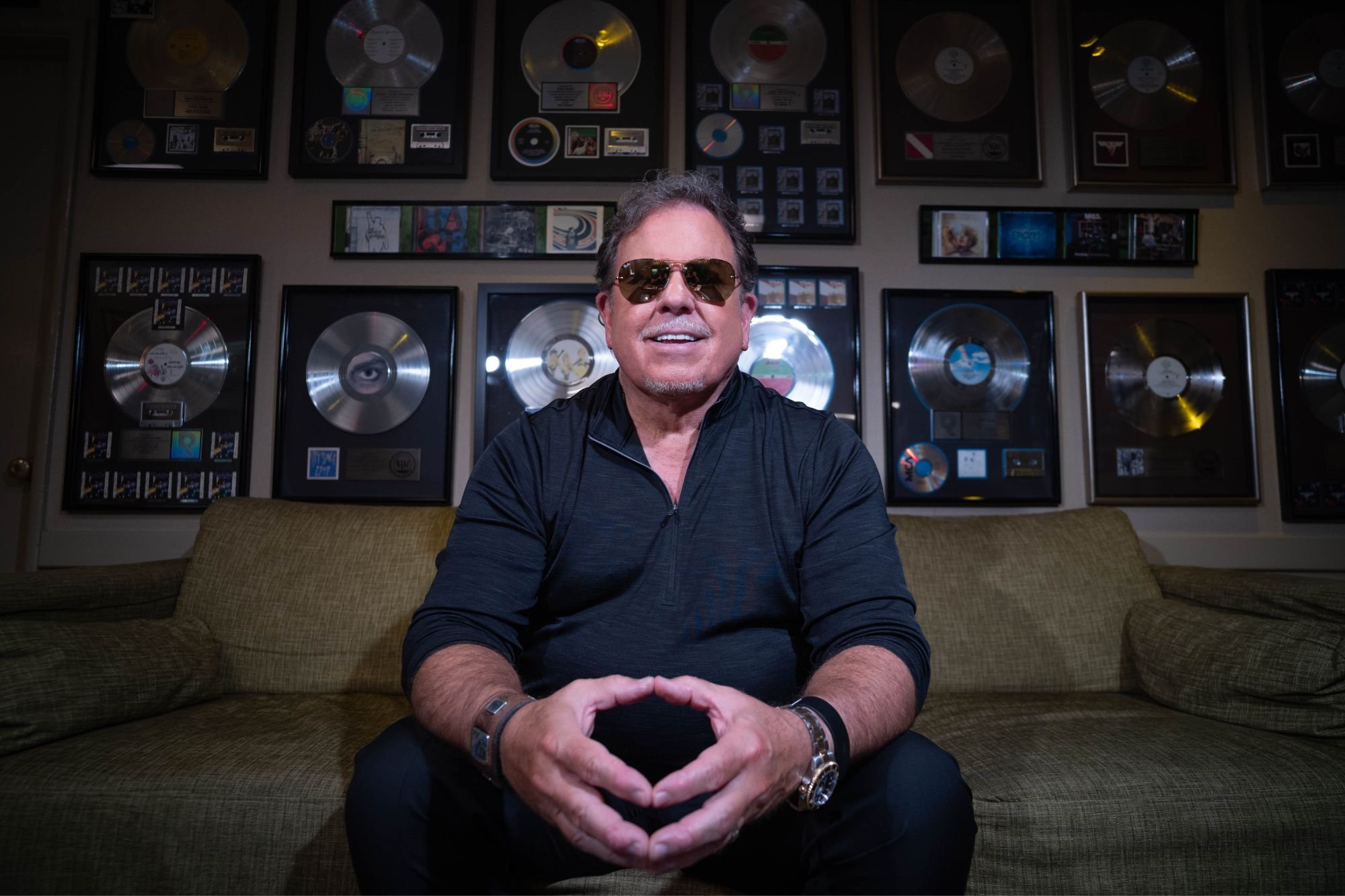 A man wearing sunglasses and a black jacket sitting on a tan sofa, smiling at the camera. The background features a wall decorated with numerous platinum and gold records framed and displayed.