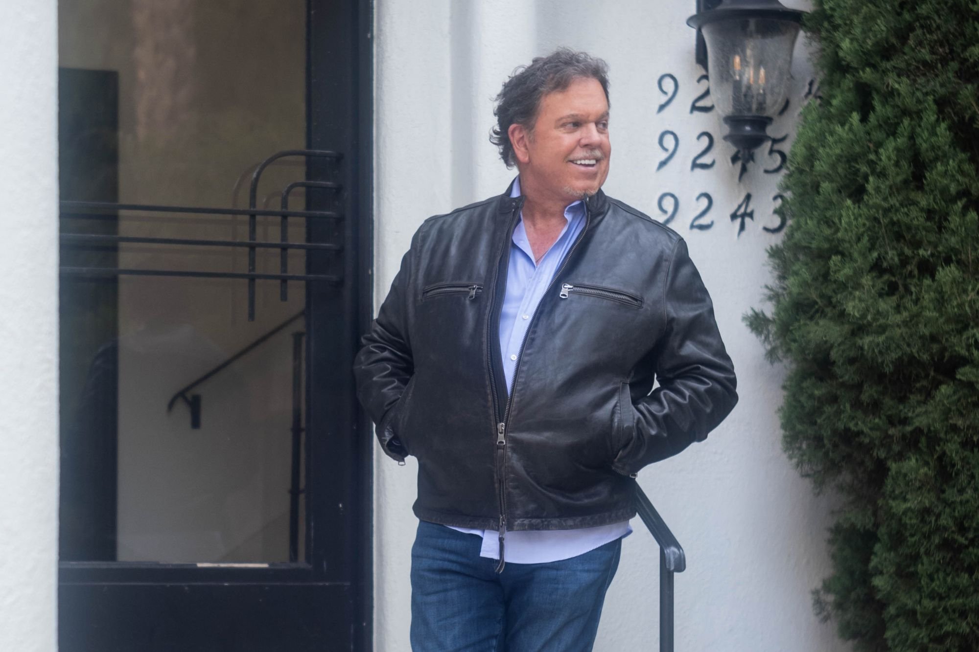 A man in a black leather jacket and blue shirt standing outside near a white building with house numbers and a lantern, smiling.