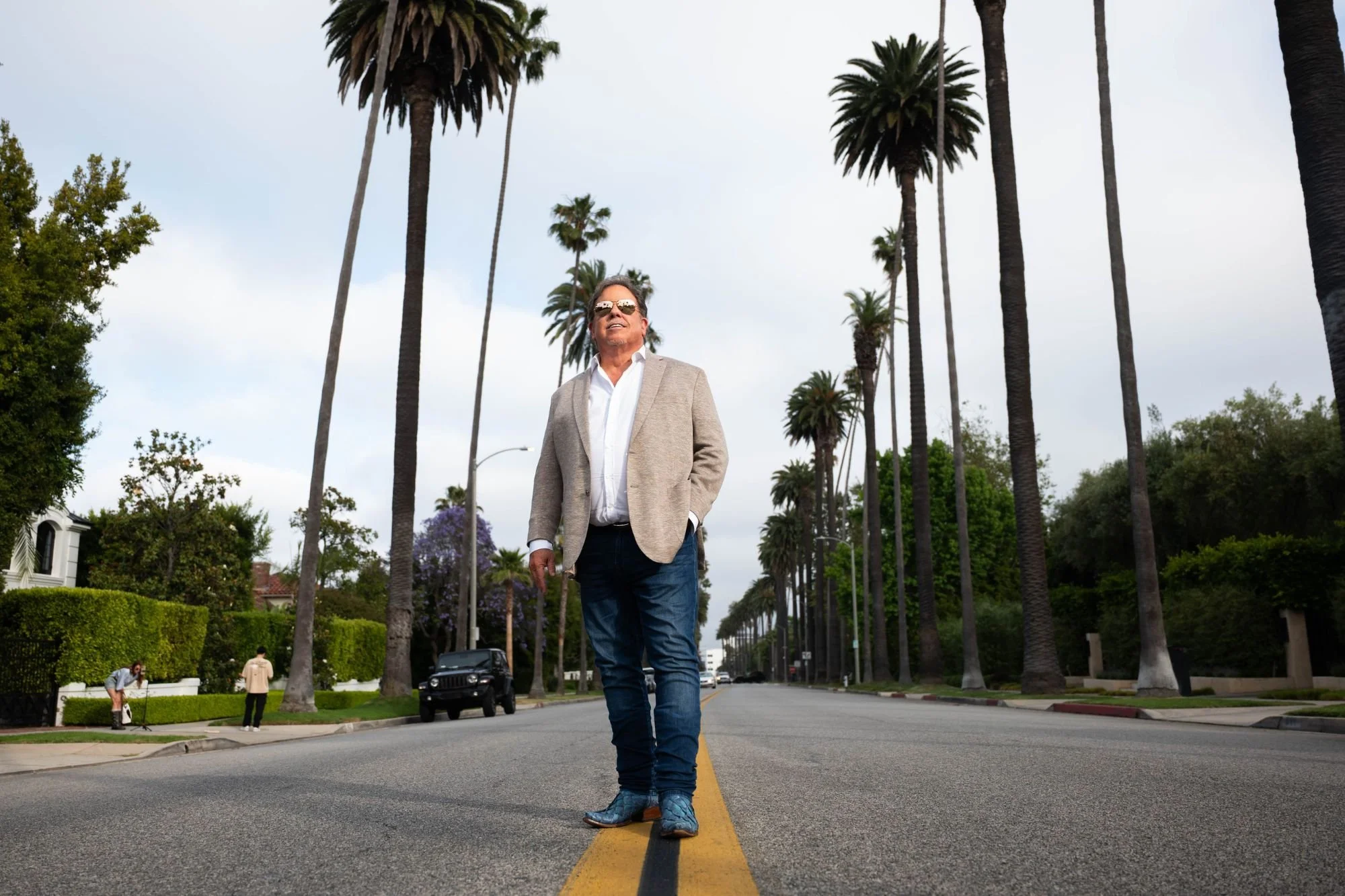 A man wearing sunglasses, a beige blazer, a white shirt, and jeans walking down the middle of a street lined with tall palm trees, with a cloudy sky overhead.