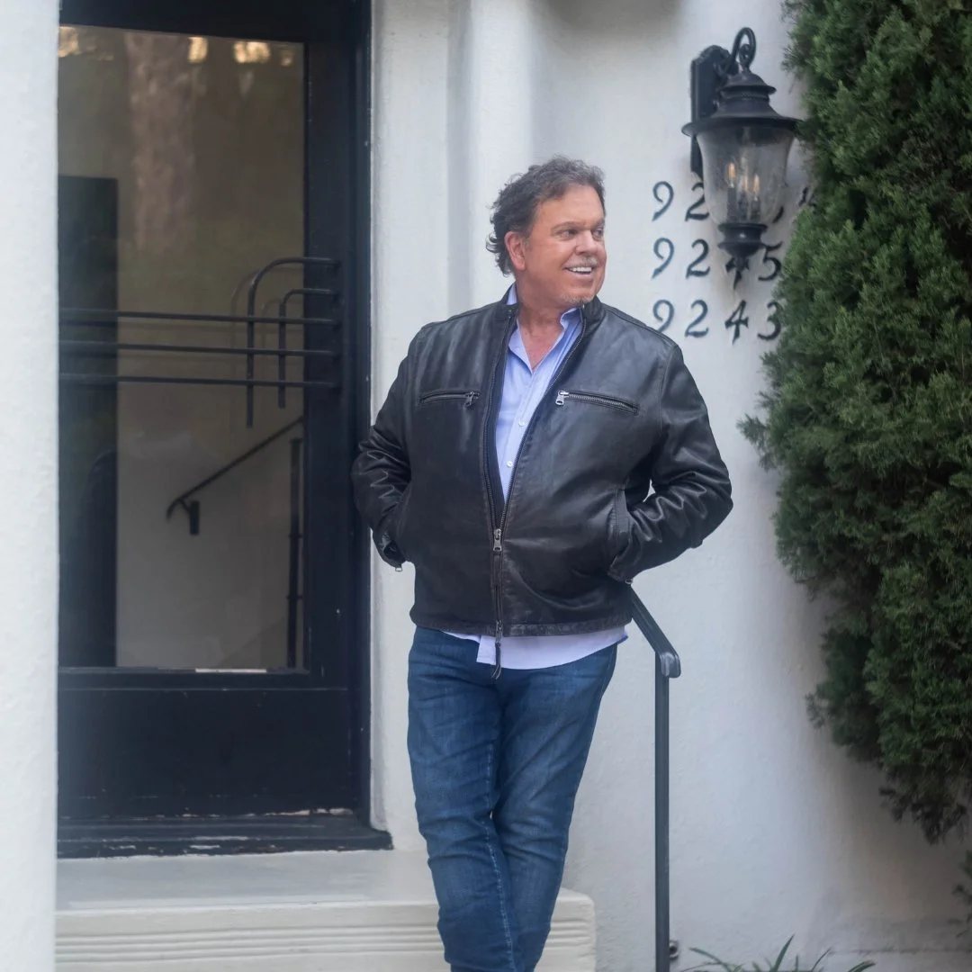 A man stands outside a house near a black door, wearing a black leather jacket and jeans, smiling and looking to the side, with house numbers and a lamp post visible on the wall beside him.