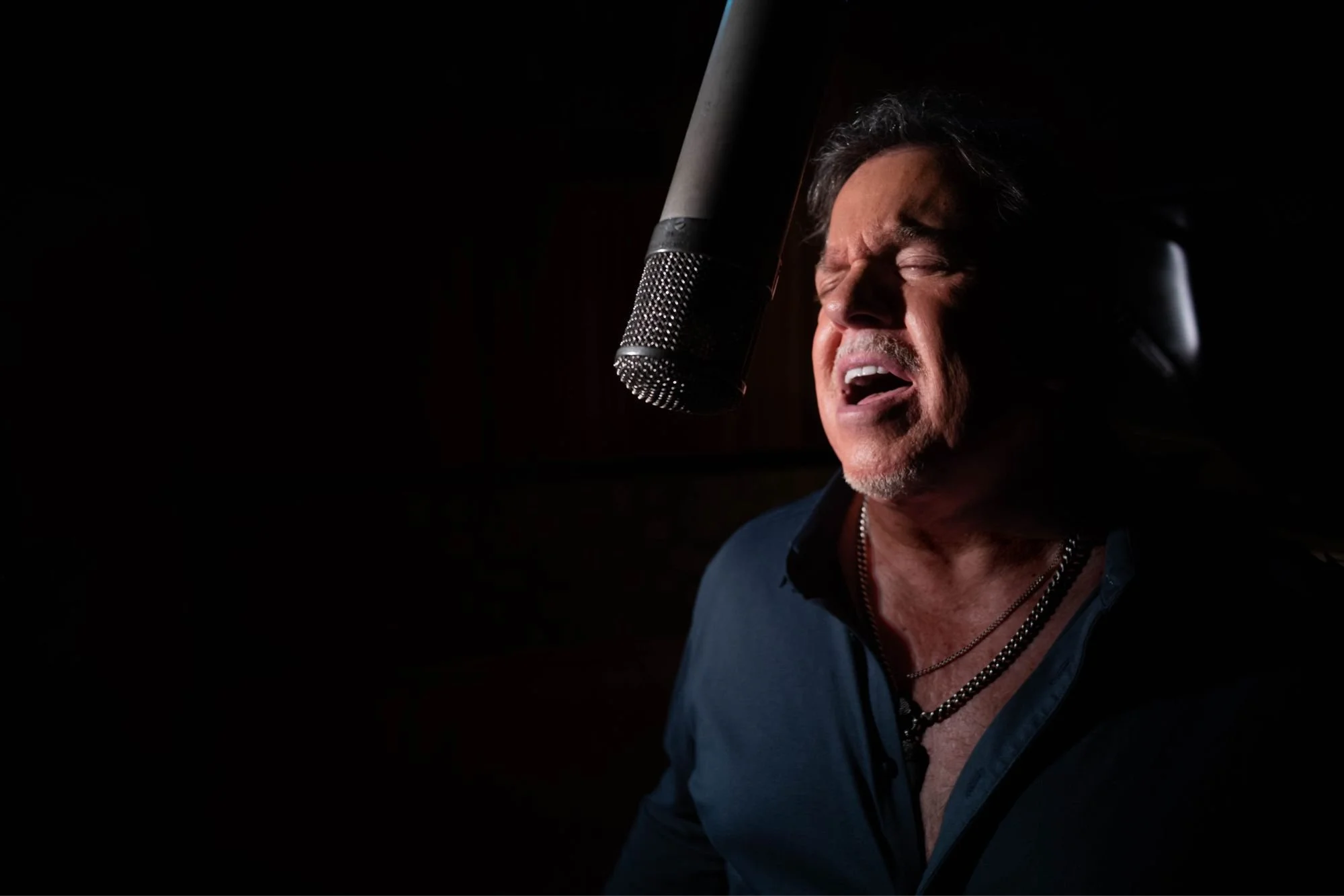 A man singing passionately into a microphone in a recording studio, with closed eyes and an emotional expression.